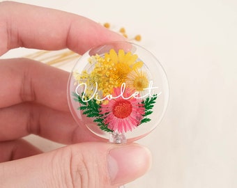 May include: A clear round badge holder with a floral design. The holder features pressed flowers in shades of yellow, pink, and green. The name "Violet" is written in white script on the front of the holder.