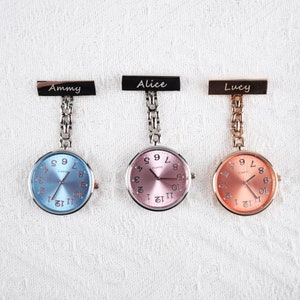 Könnte beinhalten: Drei silberne und goldfarbene Taschenuhren mit personalisierten Namensschildern. Die Uhren haben blaue, rosa und goldene Zifferblätter mit schwarzen römischen Ziffern.