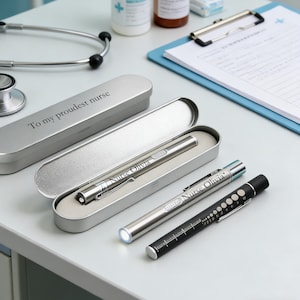May include: A collection of medical penlights and a stethoscope on a light gray surface. One penlight is in a silver case with the inscription "To my proudest nurse." Two penlights are silver, and one is black. The penlights have the name "Nurse Olivia" engraved on them.
