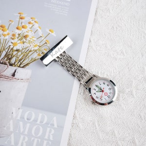May include: A silver nurse's watch with a white face and red accents. The watch has a clip on the back and a name tag that says "Kate".