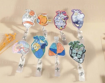 May include: A collection of medical-themed badge reels featuring various anatomical designs. The designs include a heart with a rose, an eye, a heart with a sword, a brain, a heart with a portrait, lungs, and a heart with a butterfly. Each reel has a clear plastic clip.