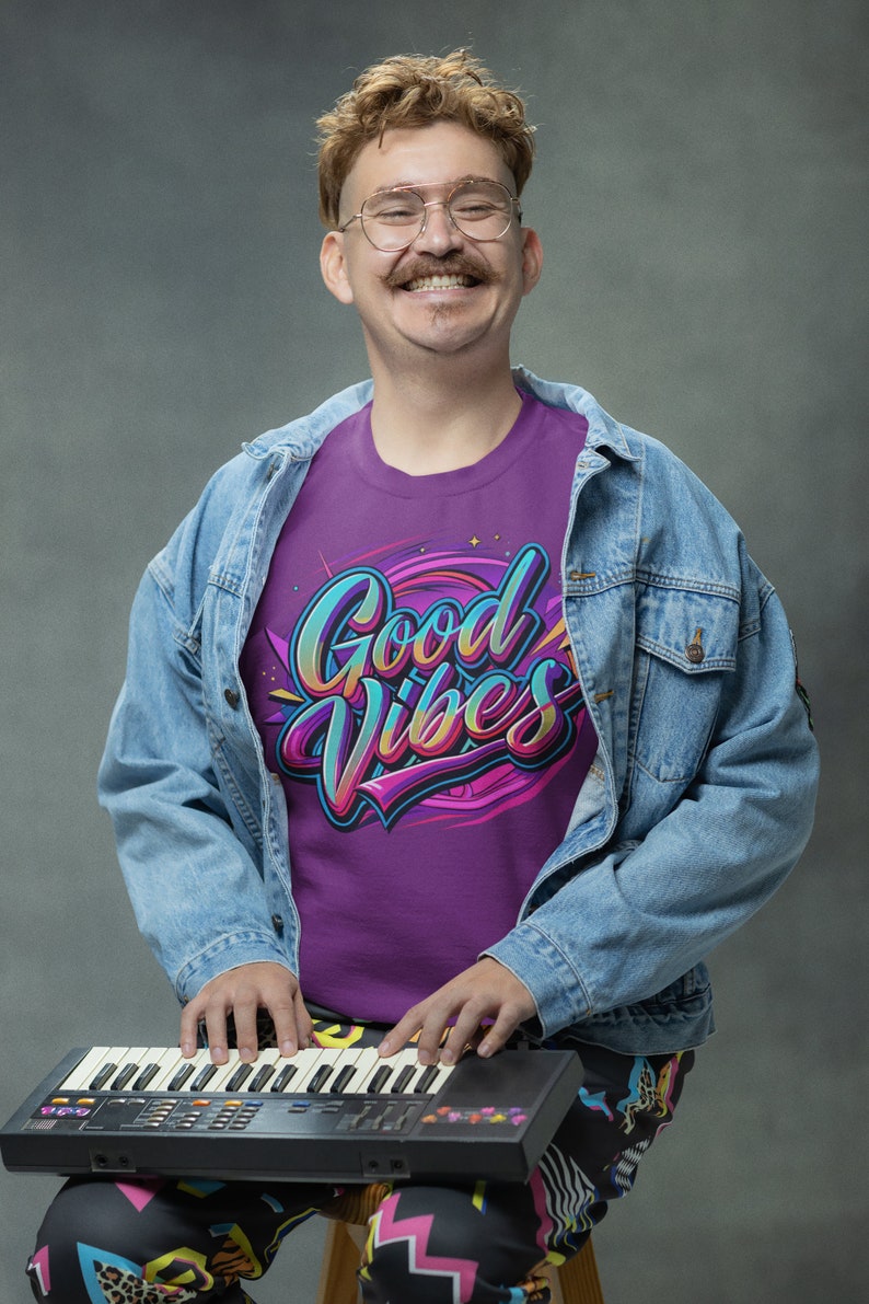 May include: A person wearing a purple t-shirt with the text "Good Vibes" in colorful lettering, a blue denim jacket, and colorful patterned pants, playing a black keyboard.