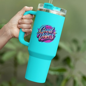 May include: A turquoise travel mug with a handle and a straw. The mug has a stainless steel lid and a graphic that says "Good Vibes" in colorful letters.