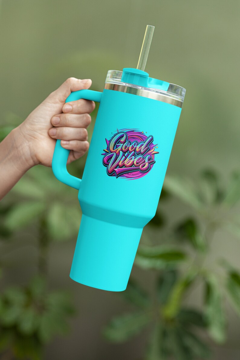 May include: A turquoise travel mug with a handle and a straw. The mug has a stainless steel lid and a graphic that says "Good Vibes" in colorful letters.
