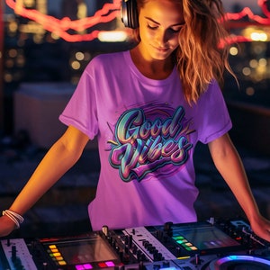 May include: A woman wearing headphones and a purple t-shirt with the text "Good Vibes" in colorful lettering, standing behind a DJ mixer with glowing lights.