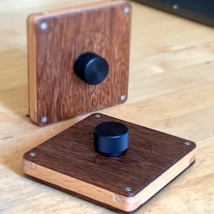 Puede incluir: Dos pomos de madera con detalles en metal negro. Los pomos son cuadrados y tienen un acabado en madera marrón oscuro con un borde de madera más claro. Los pomos están sobre una superficie de madera clara.