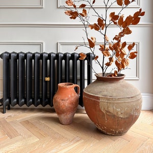 May include: A black cast iron radiator with two terracotta pots, one with a dried branch with brown leaves, and the other with a single handle, sitting on a light wood herringbone patterned floor.