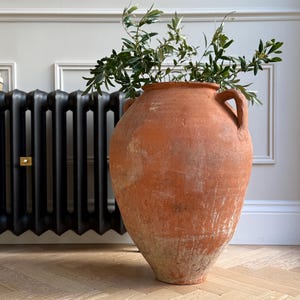 May include: A large, weathered terracotta vase with two handles, filled with green leafy branches. The vase has a wide body and a narrow neck, showing signs of age and use. The vase is placed on a light-coloured wooden floor.