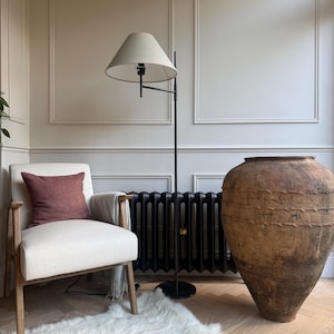 May include: An interior shot showcasing a cream armchair with a burgundy cushion, a black floor lamp with a beige lampshade, and a large, textured brown vase. A white faux fur rug and a black radiator are also visible. The room has a neutral colour palette.