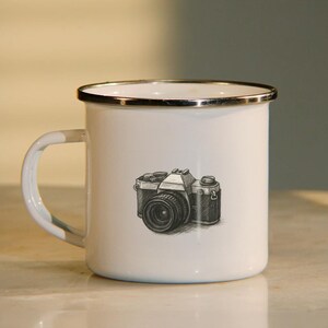 May include: White enamel mug with a silver rim featuring a black and white illustration of a vintage camera.