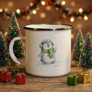 Op de afbeelding: Witte emaille mok met een zilveren rand en handvat. De mok heeft een aquarel illustratie van een pinguïn met groene oorbeschermers en een sjaal. De mok is omringd door miniatuur kerstbomen en ingepakte cadeaus.