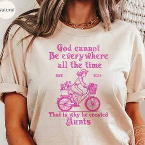 以下が含まれることがあります： ピンクのグラフィックデザインが施されたベージュのTシャツ。"神は常にどこにもいられない EST. 2024 だから彼は叔母を作った"と書かれている。グラフィックには、花かごを持った自転車に乗った女性が描かれている。