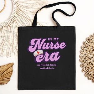 Może przedstawiać: Czarna torba typu tote z różowo-fioletowym nadrukiem z napisem "In My Nurse Era" z małym czerwonym krzyżem na białym kapeluszu. Tekst pod nadrukiem brzmi "My friends & family medical Go-to."