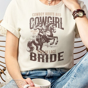 K&ouml;nnte beinhalten: Cremefarbenes T-Shirt mit brauner Grafik einer Cowgirl auf einem Pferd, Text "Cowboy Boots and Cowgirl Bride" und "September 2024". Das Shirt ist unisex.