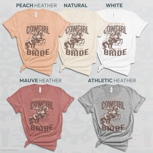 K&ouml;nnte beinhalten: Verschiedene T-Shirts in Farben wie Pfirsich, Creme, Wei&szlig;, Malve und Grau. Jedes Shirt zeigt eine Grafik einer Cowgirl auf einem Pferd mit dem Text "Cowboy Boots and Cowgirl Lace Bride."
