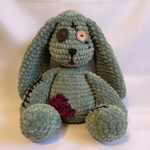 Puede incluir: Un conejo de peluche hecho a mano en un suave color verde salvia. El conejo tiene ojos de botón, detalles cosidos y un parche burdeos. El diseño incluye puntadas negras y una boca roja, dándole un aspecto único y artesanal.