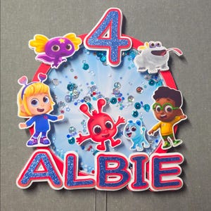 May include: A vibrant cake topper with the name "ALBIE" and the number "4" in blue glitter. The topper features cartoon characters and confetti inside a red and blue frame, ideal for a birthday party.