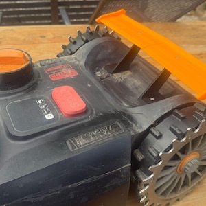 Può includere: Tosaerba robotico nero e arancione con uno spoiler arancione brillante. Il tosaerba ha grandi ruote dentate e un pannello di controllo con pulsanti. La parte superiore del tosaerba ha un tappo arancione e dettagli argentati. Il tosaerba è su una superficie di legno.