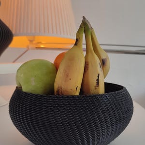 Könnte beinhalten: Eine schwarze geflochtene Schale gefüllt mit Obst, darunter Bananen und ein grüner Apfel. Die Schale hat ein strukturiertes, wellenförmiges Design.