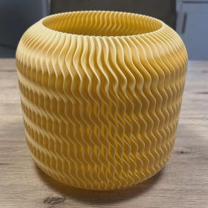 May include: A yellow, cylindrical vase with a wavy, textured design. The vase has a wide opening and a rounded base. The design creates a layered effect, adding visual interest. The vase is made of a solid material and sits on a wooden surface.