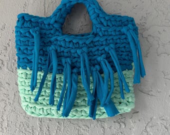 May include: A crocheted tote bag with a blue and green color scheme. The bag has a top handle and fringe details.