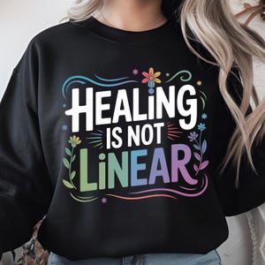 Puede incluir: Sudadera negra con el texto blanco "HEALING IS NOT LINEAR", con detalles florales y remolinos coloridos. El diseño presenta un degradado de arcoíris. Prenda casual y cómoda.
