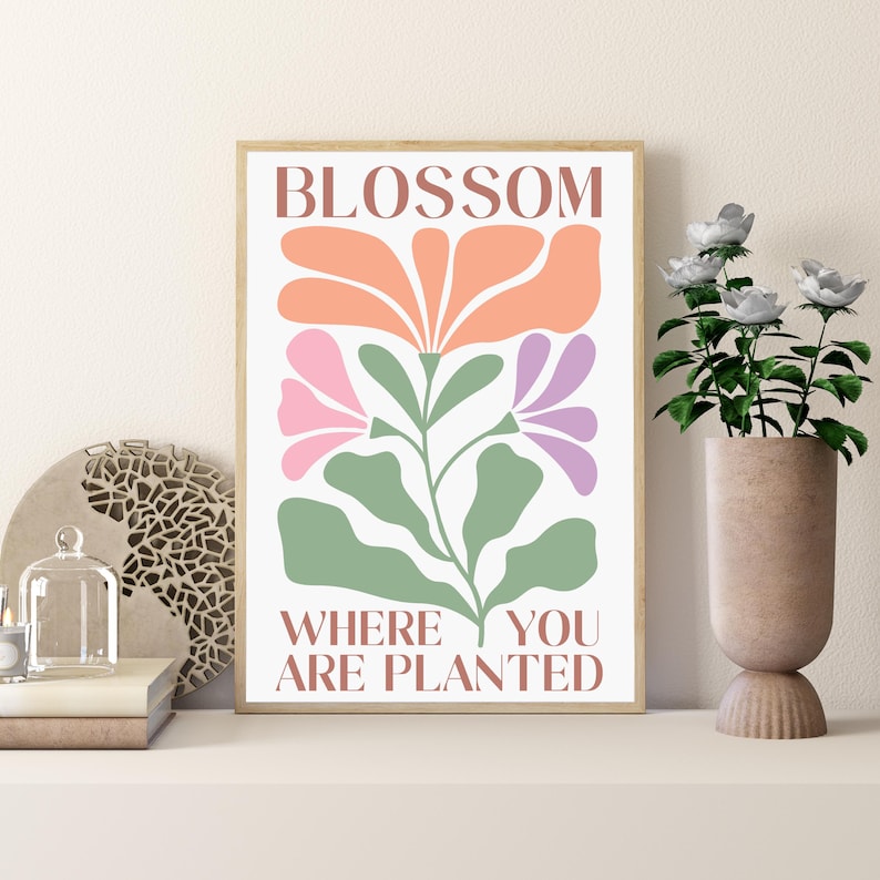 Może przedstawiać: Oprawiony wydruk artystyczny z kwiatowym wzorem w odcieniach brzoskwini, r&oacute;żu i fioletu, z zielonymi liśćmi. Tekst "BLOSSOM WHERE YOU ARE PLANTED" jest wyświetlany w kolorze brązowym. Grafika jest prezentowana na białej p&oacute;łce.