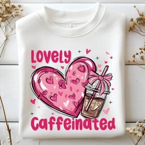 Puede incluir: Sudadera blanca con un corazón rosa y una taza de café rosa y marrón. El texto "Lovely Caffeinated" está impreso en la sudadera.