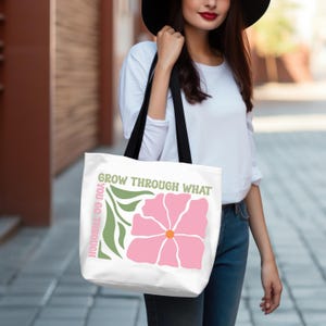 Może przedstawiać: Biała torba typu tote z r&oacute;żową grafiką kwiatową, pomarańczowym środkiem i zielonymi liśćmi. Tekst brzmi "GROW THROUGH WHAT YOU GO THROUGH" w kolorze zielonym i r&oacute;żowym. Torba ma czarne paski.
