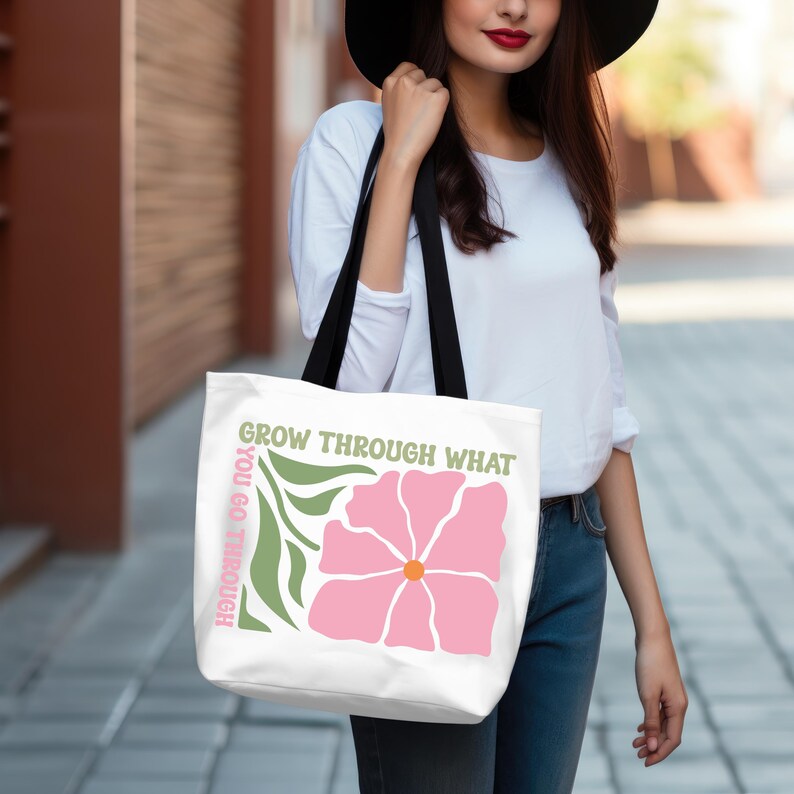 Może przedstawiać: Biała torba typu tote z r&oacute;żową grafiką kwiatową, pomarańczowym środkiem i zielonymi liśćmi. Tekst brzmi "GROW THROUGH WHAT YOU GO THROUGH" w kolorze zielonym i r&oacute;żowym. Torba ma czarne paski.