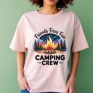 Peut inclure: T-shirt rose clair avec un motif sur le thème du camping. Le motif représente un feu de camp avec un ciel étoilé, des pins et les mots "Friends Fires Fun" et "Camping Crew" en noir.