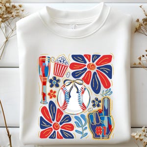 Peut inclure: Sweat-shirt blanc avec un motif sur le thème du baseball. Le graphisme comprend une balle de baseball, des fleurs, une batte, du pop-corn et un doigt en mousse. Le design présente des accents rouges, blancs, bleus et dorés.