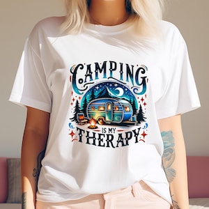 Peut inclure: T-shirt blanc avec un motif graphique coloré représentant un camping-car, un feu de camp et des arbres. Le texte "Camping is my therapy" est écrit dans une police de caractères stylisée.
