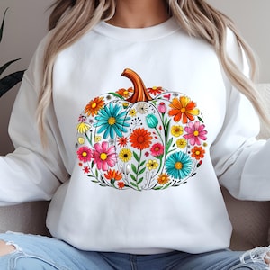 Peut inclure: Sweat-shirt blanc orné d'un motif de citrouille florale. La citrouille est remplie de fleurs colorées dans les tons de rose, orange, jaune et bleu, avec une tige marron. Le motif est centré sur le devant.