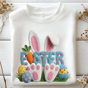Peut inclure: Sweat-shirt blanc avec un motif de Pâques coloré. Le motif comprend des oreilles de lapin, le mot "EASTER" en lettres bleues avec des fleurs roses, une carotte, des œufs de Pâques, des pattes de lapin et un poussin sortant d'un œuf.