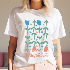 Może przedstawiać: Biała koszulka z kwiatowym wzorem. Wz&oacute;r przedstawia niebieskie i koralowe kwiaty z zielonymi łodygami i liśćmi. Słowa "Radiate Like Wildflowers" są wydrukowane w kolorze koralowym.