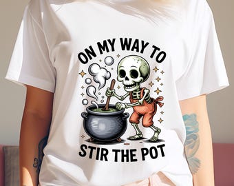 Puede incluir: Camiseta blanca con un esqueleto de dibujos animados removiendo un caldero. El esqueleto lleva un mono naranja y el texto dice "ON MY WAY TO STIR THE POT". El diseño incluye estrellas y vapor.