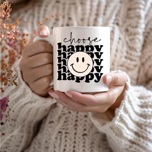 May include: White ceramic mug with black text that reads "choose happy" and a black and yellow smiley face. The mug is being held by a person wearing a white sweater.