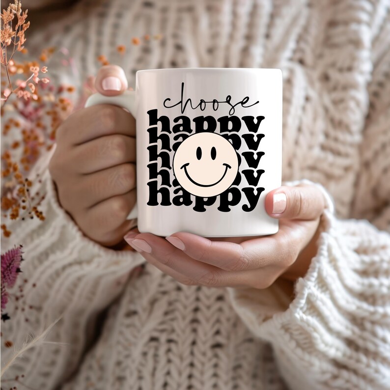 May include: White ceramic mug with black text that reads "choose happy" and a black and yellow smiley face. The mug is being held by a person wearing a white sweater.