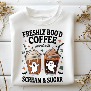 Peut inclure: Sweat-shirt blanc avec un motif sur le thème d'Halloween. Le motif présente deux tasses de café avec des illustrations de fantômes, de la crème fouettée et des pailles. Le texte dit "Freshly Boo'd Coffee Served with Scream & Sugar."