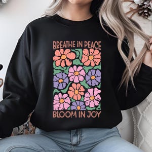 Może przedstawiać: Czarna bluza z kwiatowym wzorem w odcieniach r&oacute;żu, pomarańczy, fioletu i zieleni. Napisy "Breathe in Peace" i "Bloom in Joy" są wydrukowane nad i pod kompozycją kwiatową.