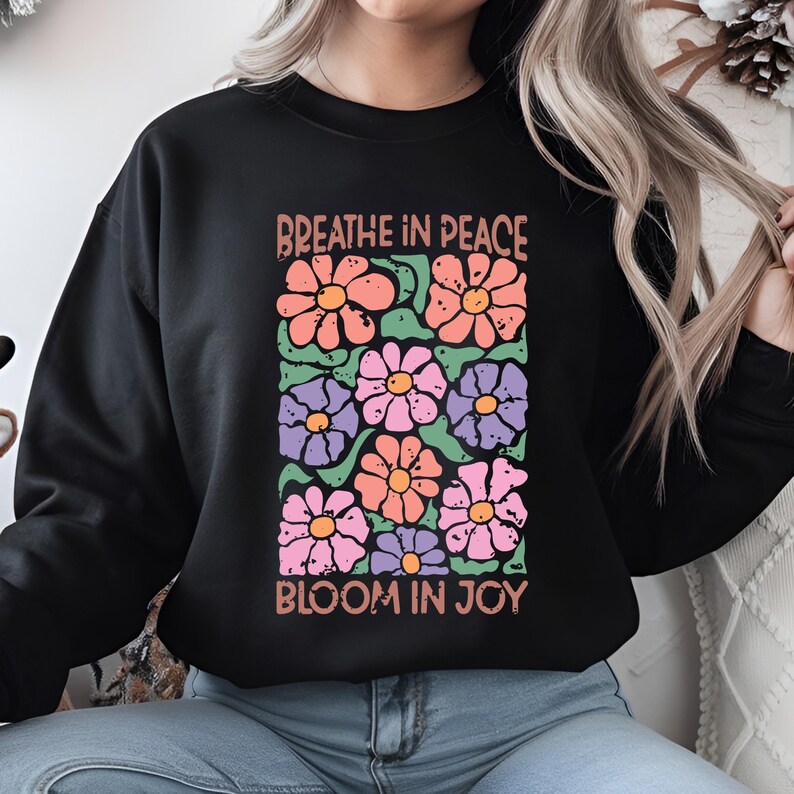 Może przedstawiać: Czarna bluza z kwiatowym wzorem w odcieniach r&oacute;żu, pomarańczy, fioletu i zieleni. Napisy "Breathe in Peace" i "Bloom in Joy" są wydrukowane nad i pod kompozycją kwiatową.