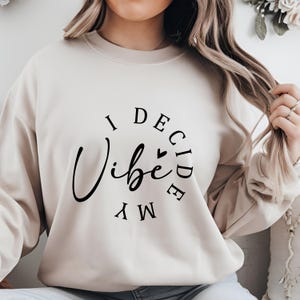 May include: A beige sweatshirt with the text "I Decide My Vibe" in black lettering.