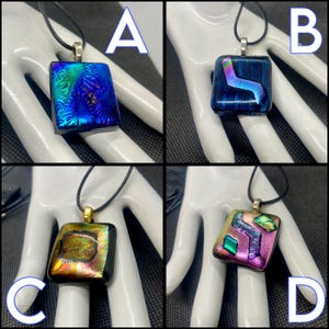 May include: Four square dichroic glass pendants on black cords. Each pendant has a different design and color scheme. The pendants are displayed on a white hand-shaped display. The pendants are labeled A, B, C, and D.