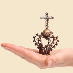 Könnte beinhalten: Eine kleine, verzierte Krone mit einem Kreuz oben drauf. Die Krone ist aus Metall und hat klare Strasssteine. Sie liegt auf der Handfläche.