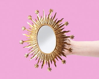 May include: A gold-colored sunburst mirror with an oval mirror in the center. The sunburst design features pointed rays and star accents. The mirror is held against a pink background.