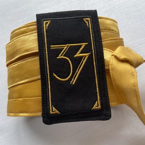 May include: A black rectangular patch embroidered with the number "33" in gold, framed by a gold border. The patch is next to a rolled-up gold ribbon and a tied gold ribbon.