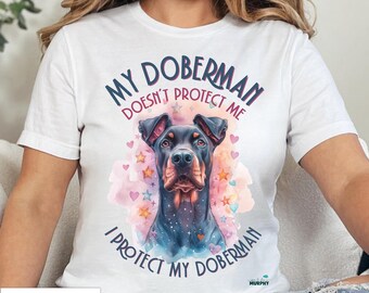 May include: White t-shirt featuring a Doberman Pinscher illustration with the text "MY DOBERMAN DOESN'T PROTECT ME I PROTECT MY DOBERMAN". The design includes a watercolor-style background with stars and hearts.