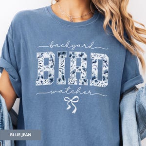 Op de afbeelding: Een blauw spijkerbroekkleurig t-shirt met de woorden "backyard bird watcher" in witte letters. Het woord "BIRD" staat in grote blokletters gevuld met een bloemen- en vogelontwerp. Een witte strik staat onder de tekst.
