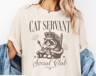 May include: Ivory-colored t-shirt featuring a regal cat illustration wearing a crown and holding a scepter. The text "CAT SERVANT" is above the cat, with "Social Club" and "Proud and Loyal" below. The shirt has a relaxed fit.